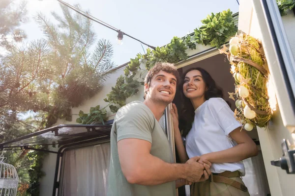 Low angle view of smiling couple holding hands near door of camper van outdoors — Photo de stock