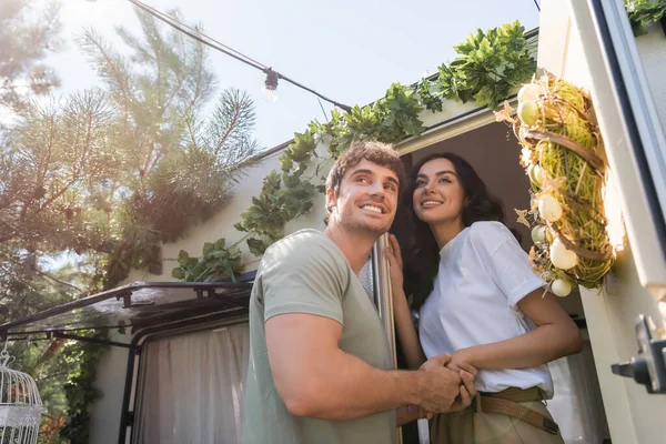 Low angle view of smiling couple holding hands near door of camper van outdoors - foto de stock