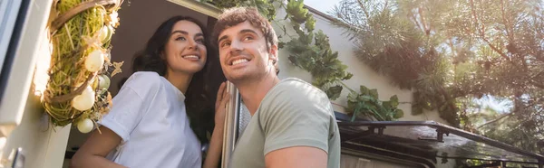 Low angle view of cheerful young couple looking away near door of camper van outdoors, banner - foto de stock