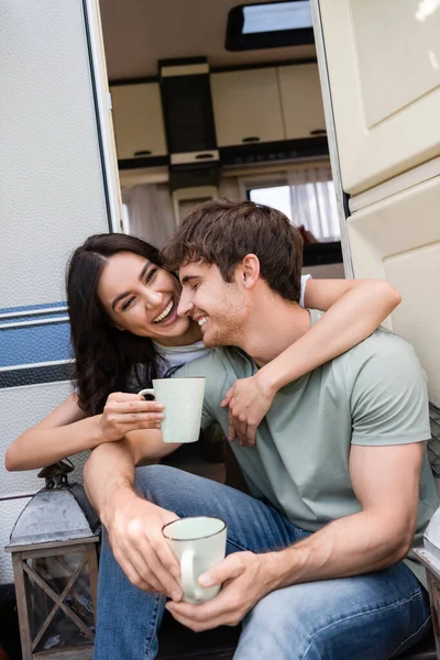 Positive young woman hugging boyfriend with cup near door of camper van — Stock Photo