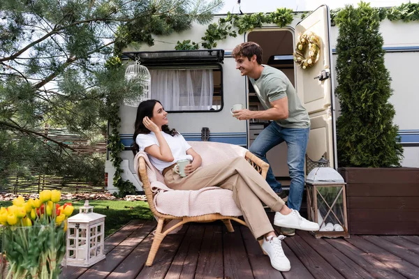 Side view of cheerful man holding cup near girlfriend on armchair and camper van — Stock Photo