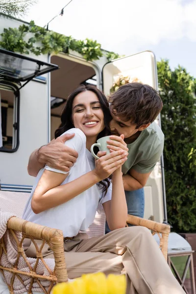 Young man kissing cheerful girlfriend with cup near camper van outdoors — Foto stock