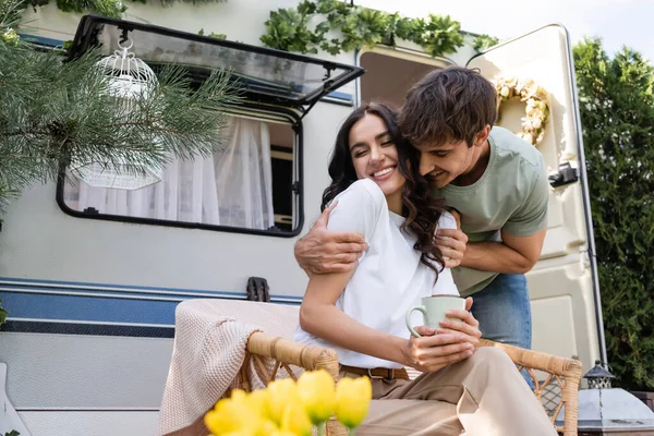 Smiling man hugging cheerful girlfriend with closed eyes holding cup near camper van — Stockfoto