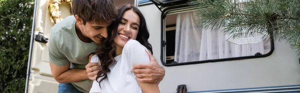 Smiling man hugging girlfriend near camper van outdoors, banner — Stockfoto