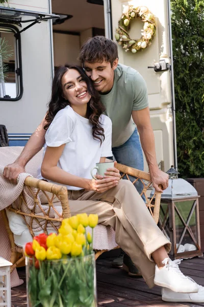 Smiling man standing near cheerful girlfriend with cup on armchair near camper van — Fotografia de Stock