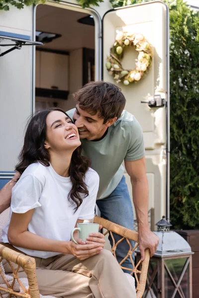 Young man kissing brunette girlfriend with cup on armchair near camper van — Stock Photo