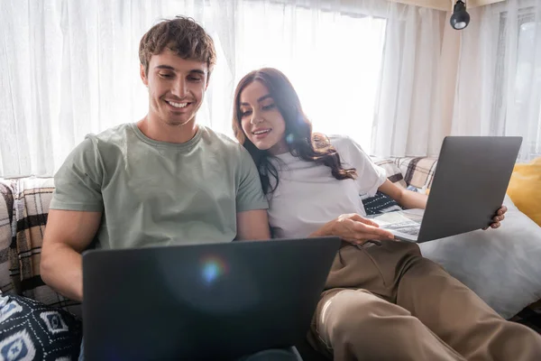Positive woman looking at laptop near blurred boyfriend on bed in camper van — Stock Photo
