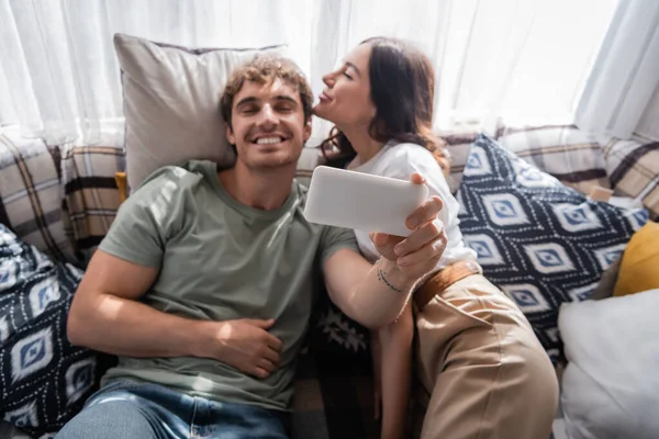 Blurred man taking selfie on smartphone near girlfriend on bed in camper van - foto de stock