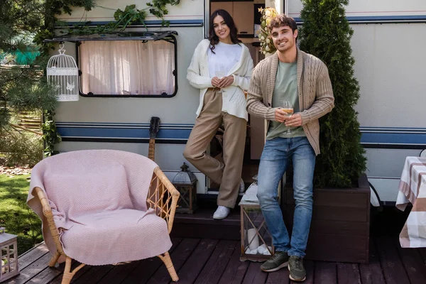 Smiling couple in cardigans holding glasses of wine near camper van — Stock Photo