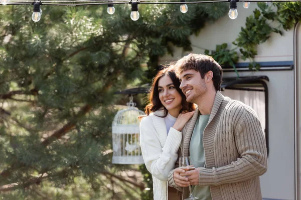 Woman in cardigan hugging boyfriend with glass of wine near blurred camper van — Foto stock