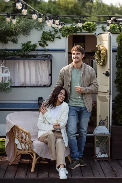 Cheerful young man holding wine and hugging girlfriend in cardigan near camper van — Fotografia de Stock