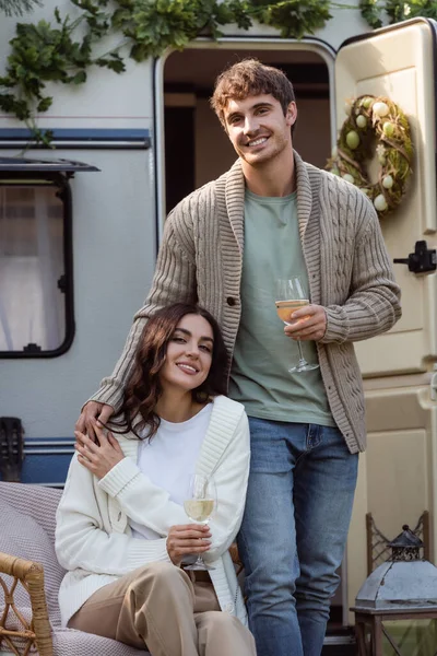 Young couple in warm cardigans holding wine and smiling at camera near camper van — Stock Photo