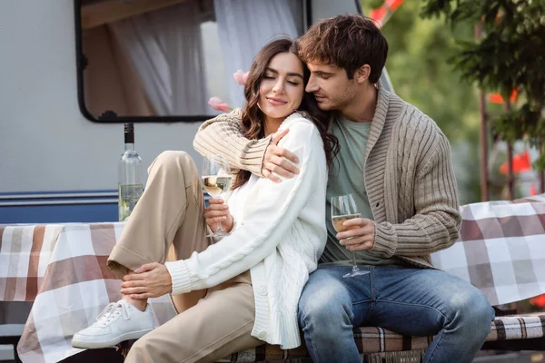 Brunette man in cardigan hugging girlfriend with wine on terrace of camper van — стоковое фото