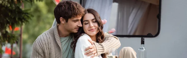 Man in knitted cardigan hugging girlfriend near glass of wine and camper van outdoors, banner — стоковое фото