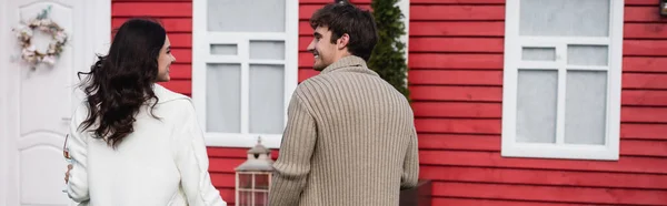 Side view of cheerful couple in knitted cardigans looking at each other near house, banner — стоковое фото