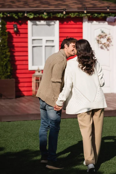 Smiling man kissing girlfriend in knitted cardigan near blurred house outdoors — Fotografia de Stock