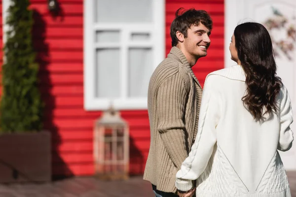 Smiling man in cardigan holding hand of brunette girlfriend near blurred house — Stock Photo