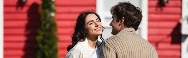 Positive couple in warm cardigans standing near blurred house outdoors, banner — Stock Photo