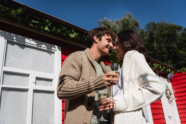 Low angle view of smiling young couple in cardigans holding glasses of wine near house outdoors — стоковое фото