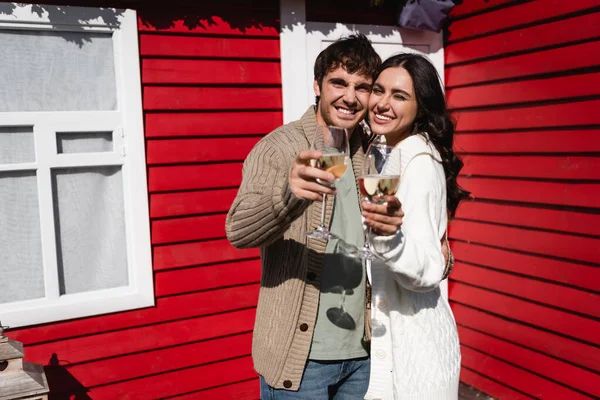 Positive couple in cardigans holding glasses of wine near house — Stock Photo