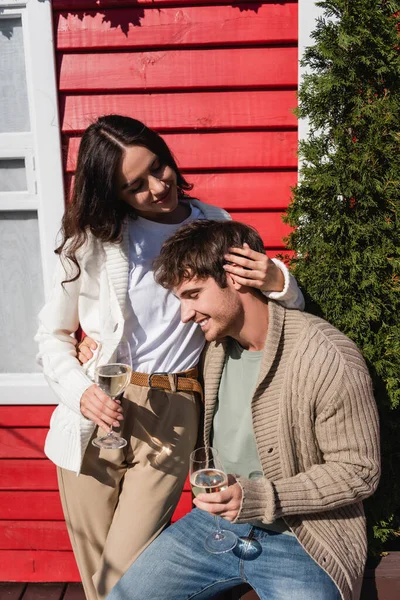 Smiling woman in cardigan hugging boyfriend with glass of wine near house — Stock Photo