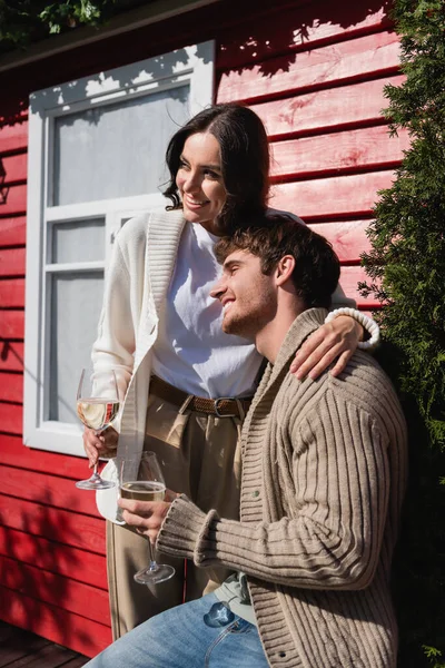 Positive couple in knitted cardigans holding glasses of wine near house - foto de stock
