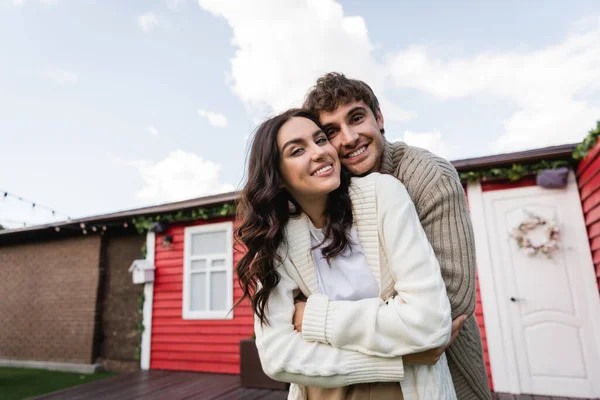 Cheerful couple in cardigans hugging and looking at camera near house outdoors — Stock Photo