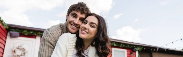 Smiling couple in knitted cardigans looking at camera near blurred house outdoors, banner — Stock Photo