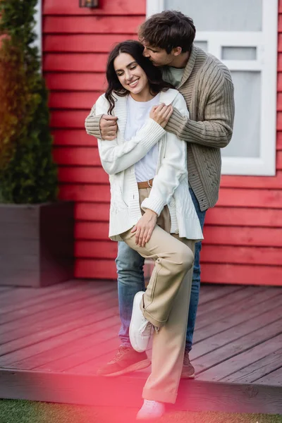 Young man kissing smiling girlfriend in knitted cardigan near house — Stockfoto