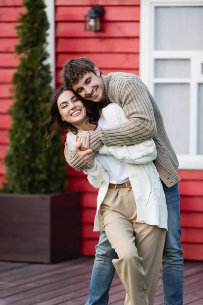 Happy man embracing girlfriend in warm cardigan near blurred house — Stock Photo