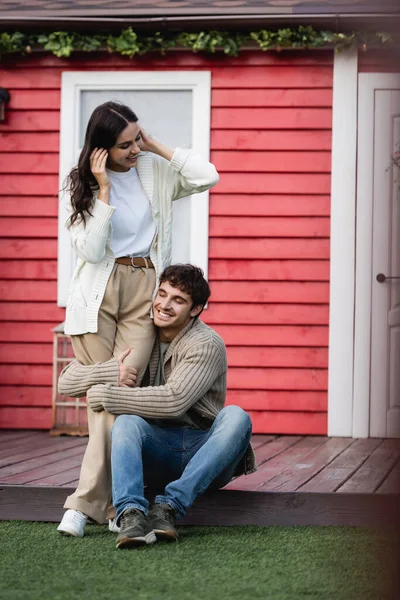 Cheerful man in cardigan hugging legs of brunette girlfriend near house — Stockfoto