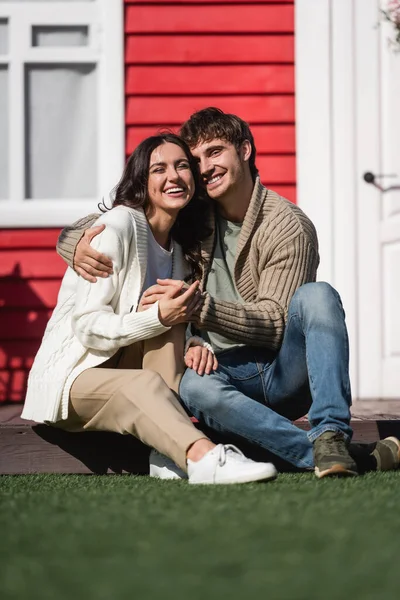 Smiling couple in cardigans hugging and looking at camera on terrace of house — Fotografia de Stock