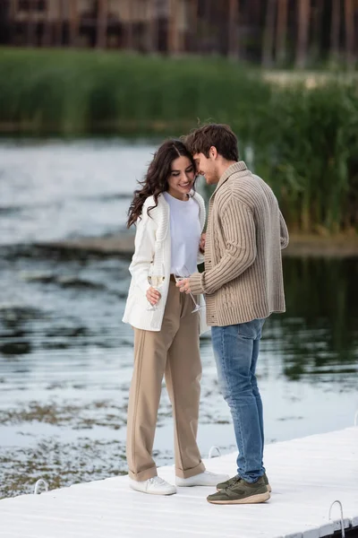 Smiling couple in cardigans holding wine on pier near river — Stock Photo