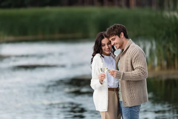 Cheerful couple in knitted cardigans toasting wine near blurred river — Stock Photo