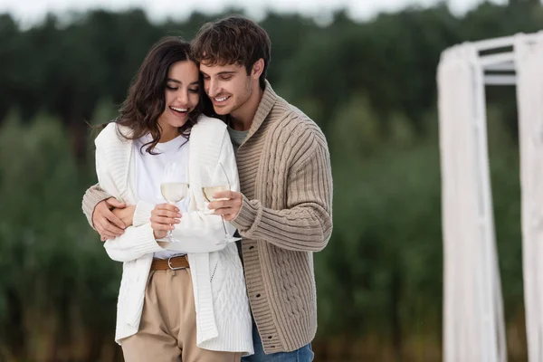 Smiling man in cardigan hugging girlfriend with wine outdoors — Stock Photo