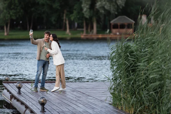 Smiling couple in cardigans holding wine on pier near lake — Stock Photo