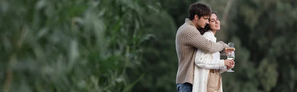 Man in cardigan hugging smiling girlfriend with wine outdoors, banner — Stock Photo