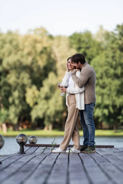 Positive man hugging girlfriend in cardigan holding wine on pier — Stock Photo