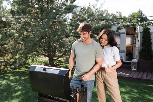 Smiling woman holding wine near boyfriend cooking on grill and camper van - foto de stock