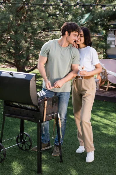 Positive woman holding wine and kissing boyfriend near grill outdoors — Stock Photo