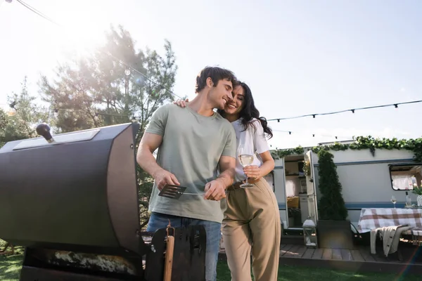 Low angle view of smiling couple holding wine and cooking near camper van — Fotografia de Stock