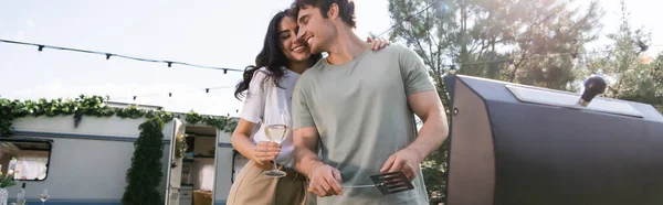 Low angle view of woman holding vine and hugging boyfriend near grill and camper van, banner — Stock Photo