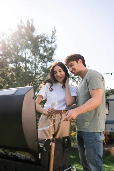 Smiling man cooking on grill near girlfriend with wine outdoors — Foto stock