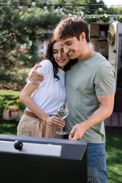 Smiling man hugging girlfriend with wine near blurred grill outdoors — Foto stock