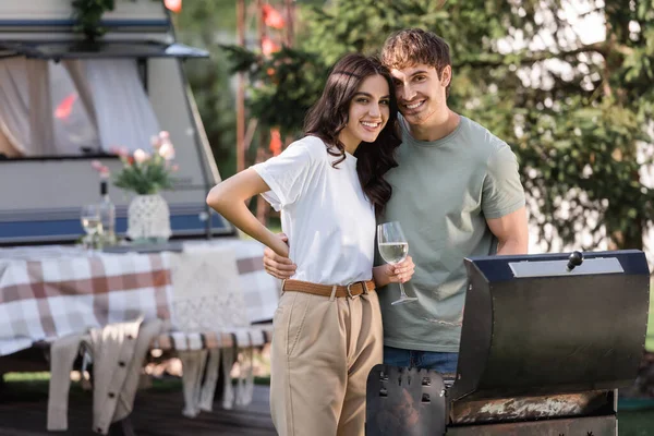 Happy couple with wine looking at camera near grill and camper van - foto de stock