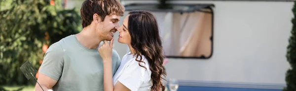 Side view of positive woman kissing boyfriend near blurred camper van, banner - foto de stock