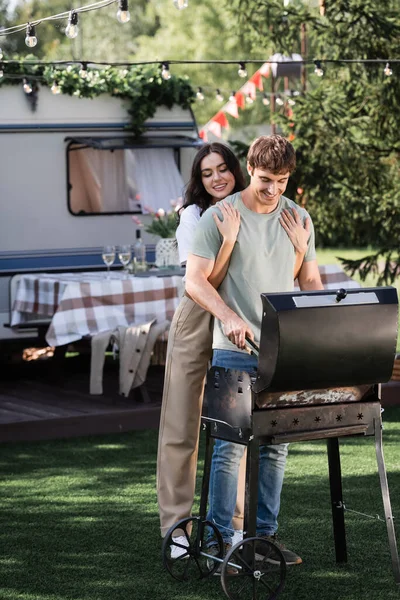 Young woman hugging boyfriend cooking on grill near blurred camper van — Stock Photo