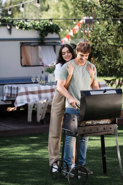Young woman hugging boyfriend cooking on grill near blurred camper van — Foto stock
