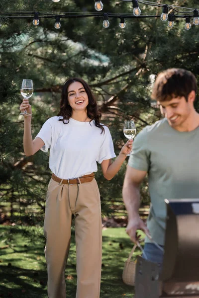 Cheerful woman holding wine near blurred boyfriend cooking on grill outdoors — Foto stock