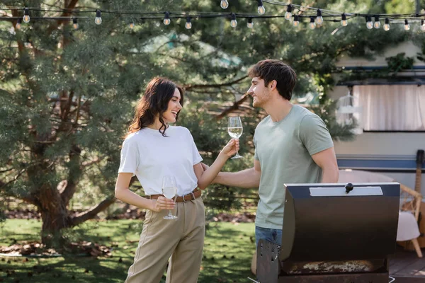 Smiling young woman holding glasses of wine near boyfriend and grill outdoors — Stock Photo